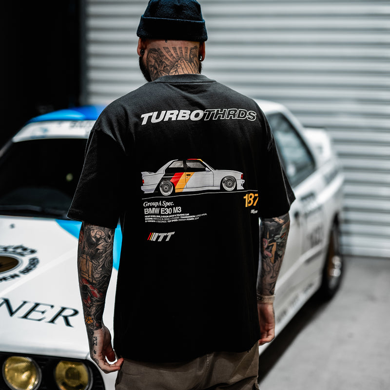 Tattooed man in front of race car wearing Turbo Threads relaxed black tee with a BMW E30 M3 graphic